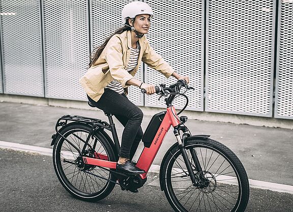 Eine Frau fährt auf einem roten Elektrorad auf Asphalt, im Hintergrund graue, gelochte Metallwände. 
